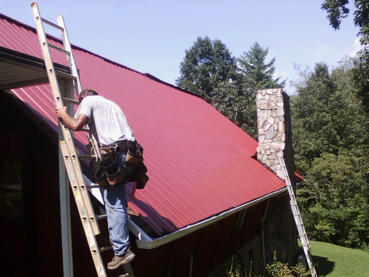 Roofer on a ladder beside a red metal roof and stone chimney — Soffit & Fascia Repair by Roofing Brothers in Phoenicia